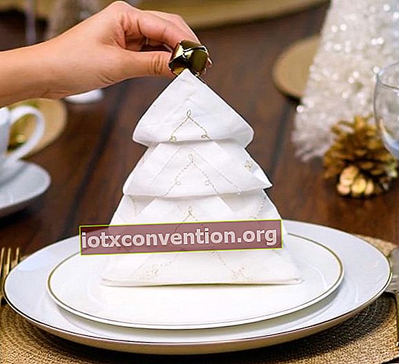 Weiße Serviette mit einer Glocke in einen Weihnachtsbaum gefaltet und auf einen weißen Teller gelegt
